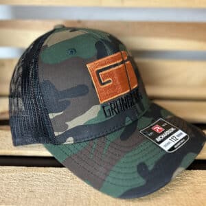 A Granberg Snap Back Trucker Mesh Hat (Camo/Black) is displayed on a wooden chair.