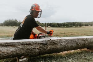A person operating a chainsaw mill to cut a log into planks in an open field, wearing safety gear including a helmet and face shield.